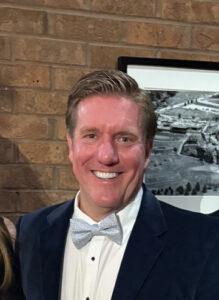 IMG_4138 1 | WellCheck Smiling man in a navy suit and light gray bow tie stands in front of a brick wall with a framed black-and-white photo behind him.