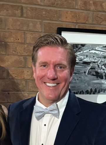 IMG_4138 1 | WellCheck Smiling man in a navy suit and light gray bow tie stands in front of a brick wall with a framed black-and-white photo behind him.